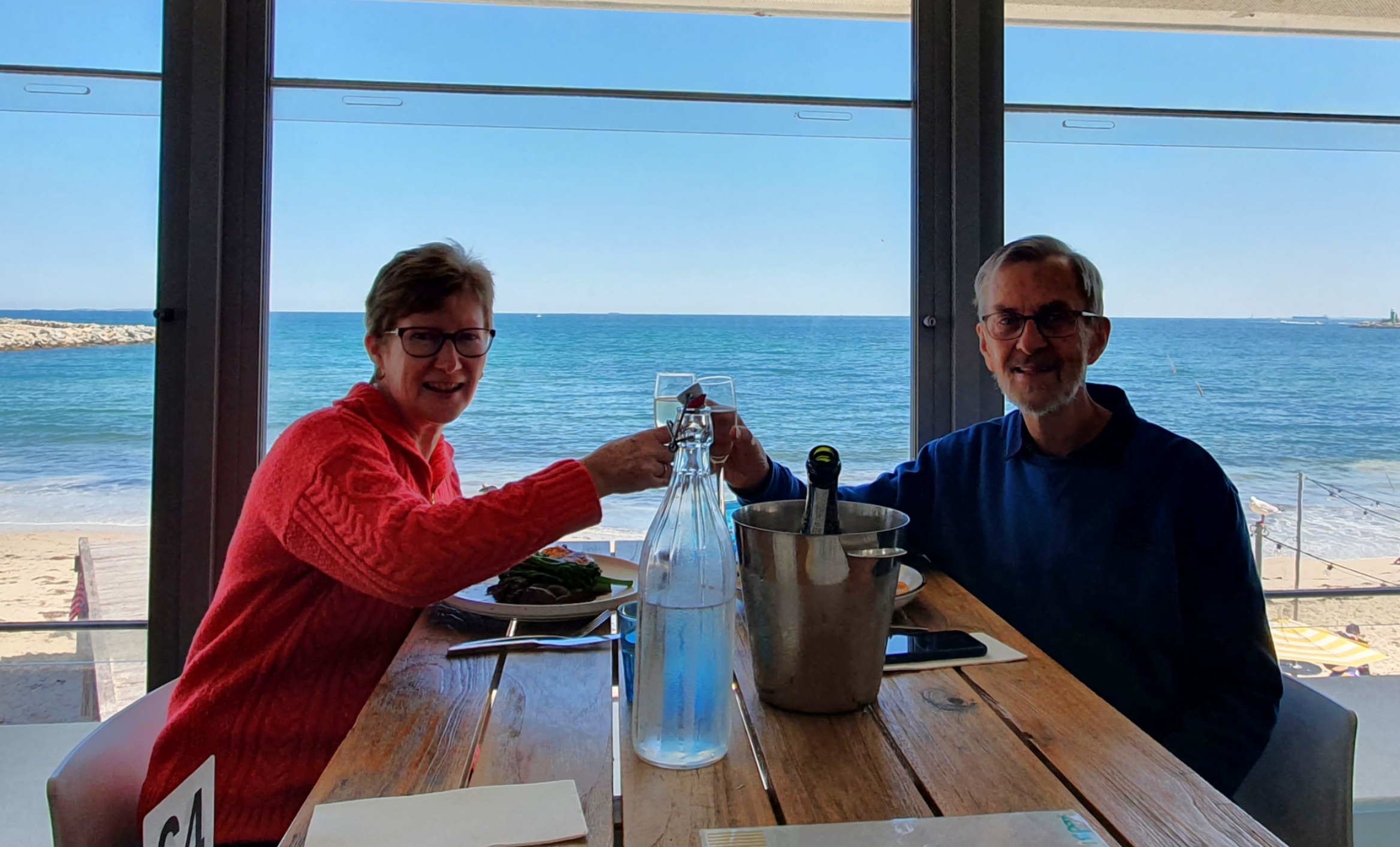 A man and a woman raising their wine glasses to each other with the ocean in the background
